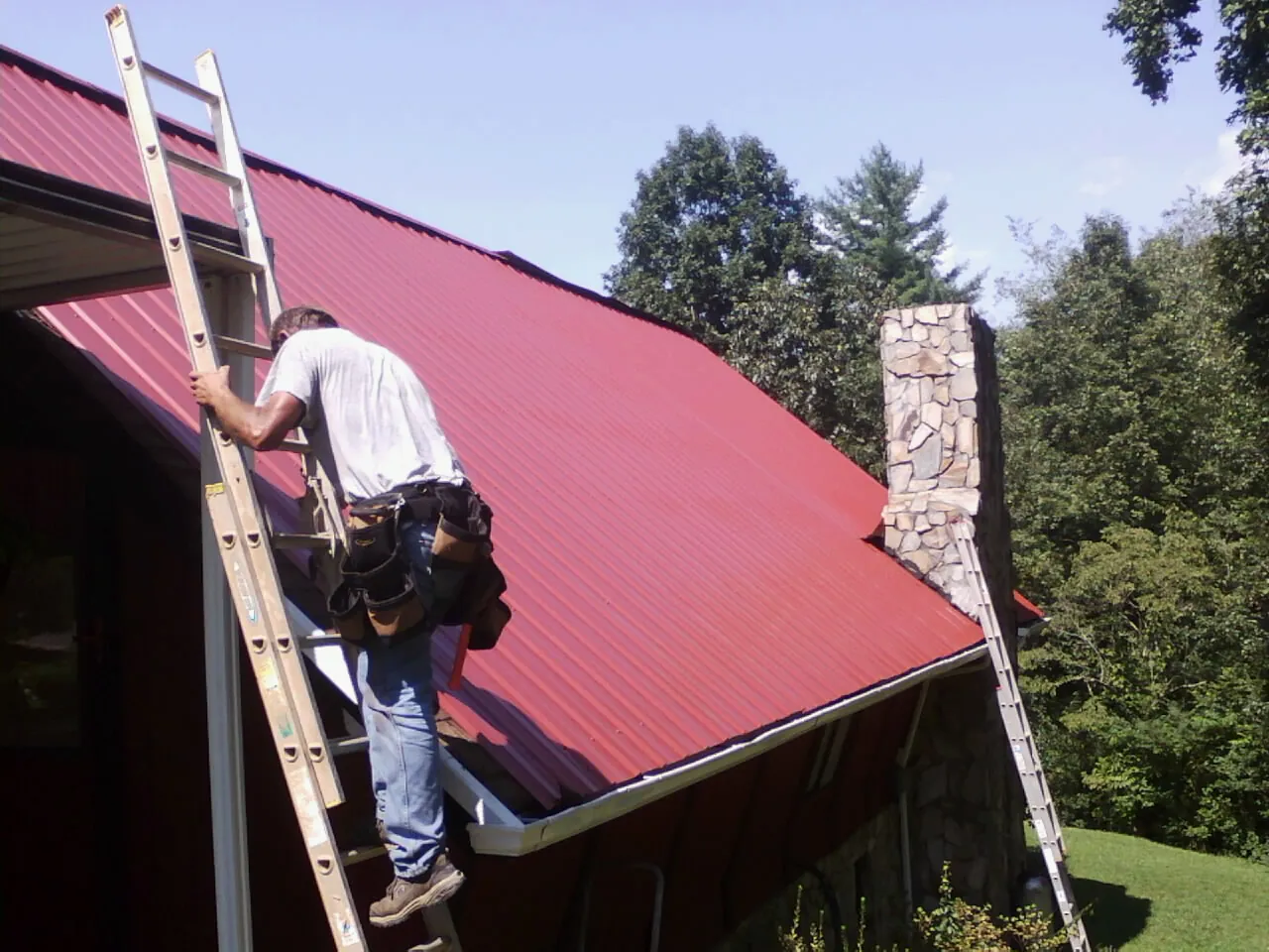 Roofer on a ladder beside a red metal roof and stone chimney — Soffit & Fascia Repair by Roofing Brothers in Auburndale